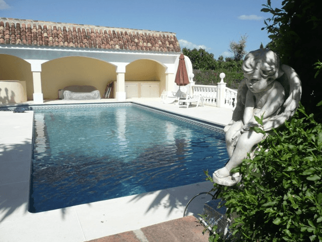 Cherub statue overlooking a clear blue swimming pool and white arched pool house.