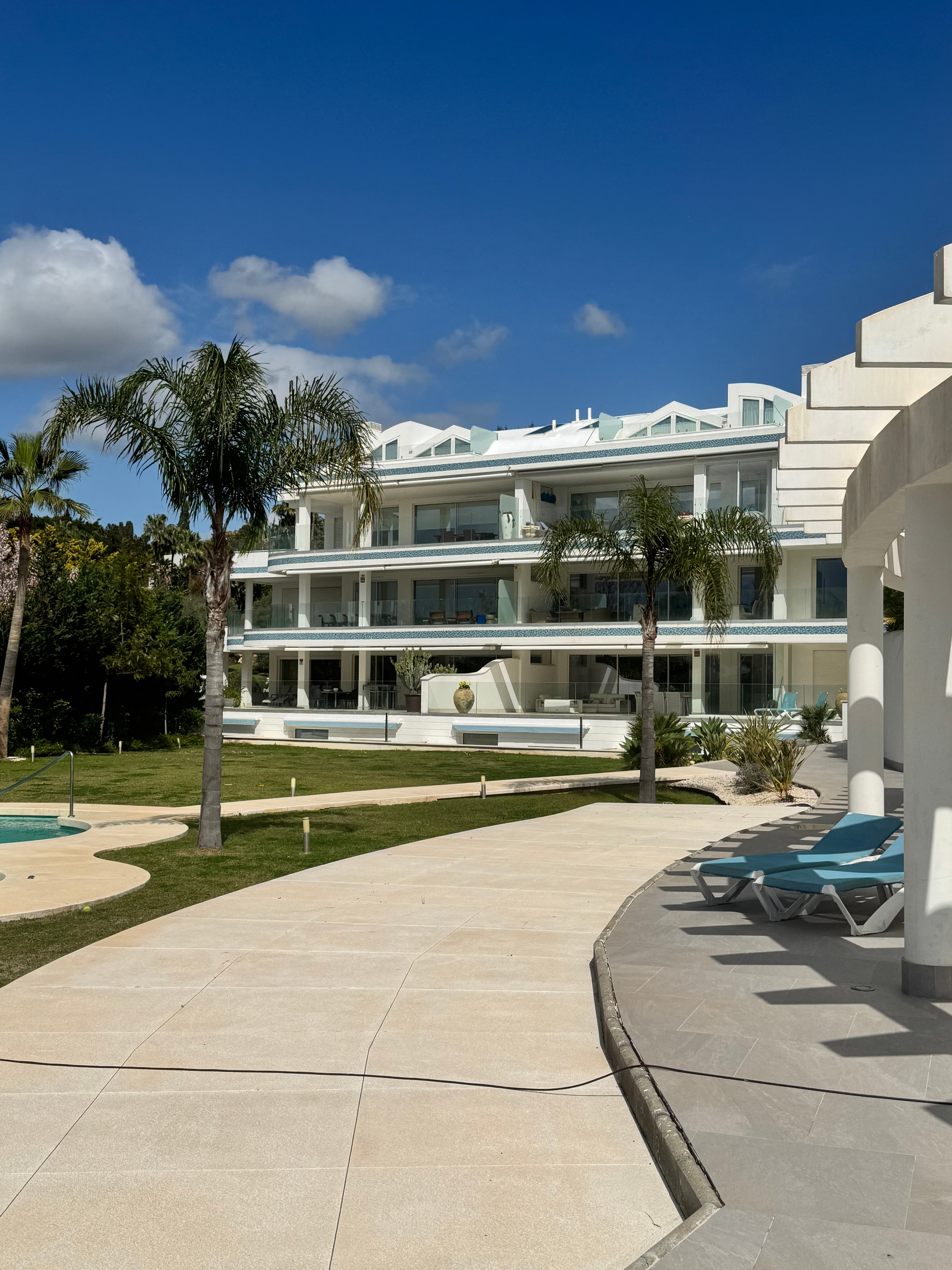 Modern white multi-story building with palm trees and a stone walkway under blue skies.