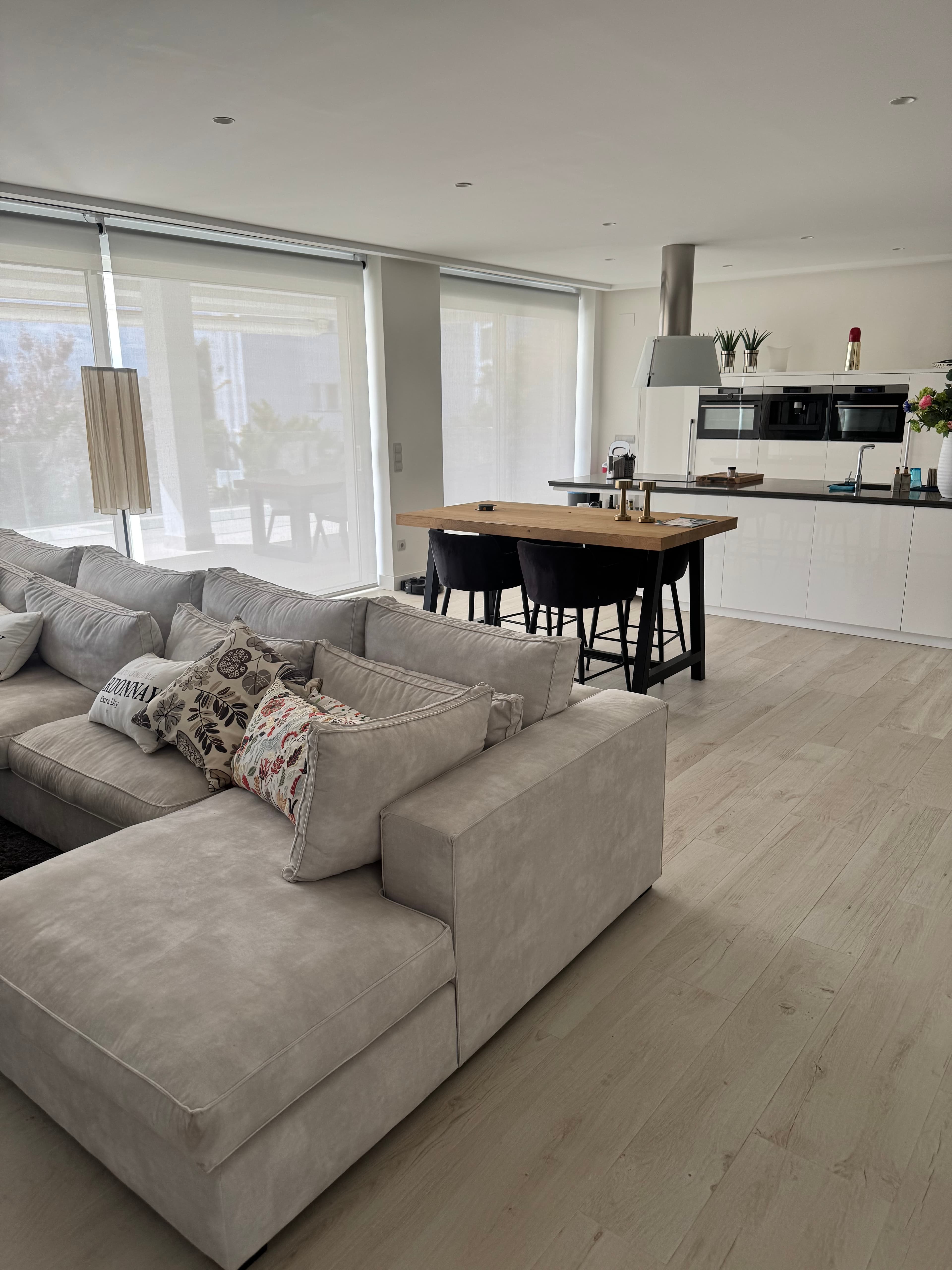 Modern living area with a gray sectional sofa, wooden dining table, and white kitchen.