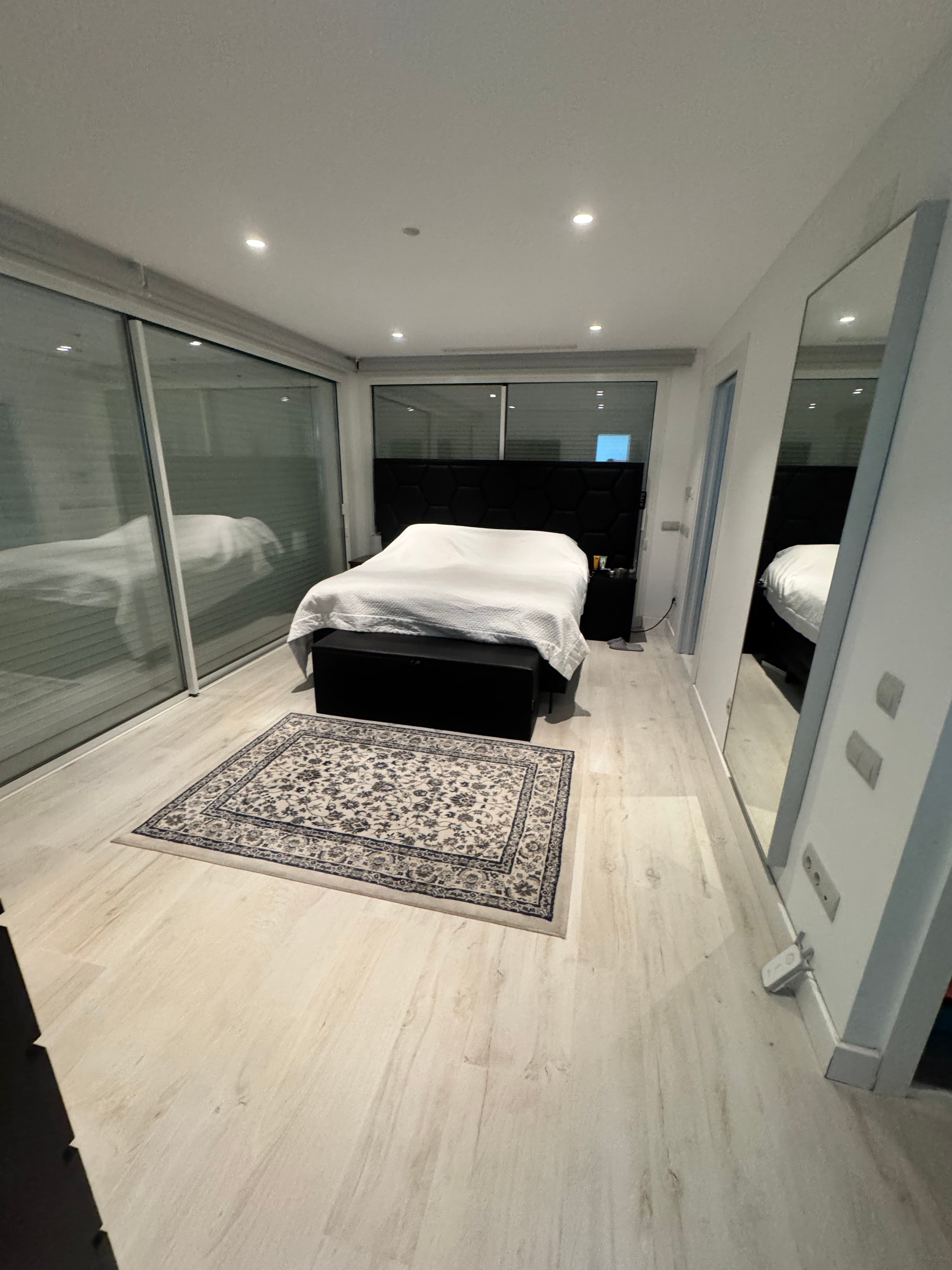 Modern bedroom with a white bed, patterned rug, light wood floors, and large mirrors.
