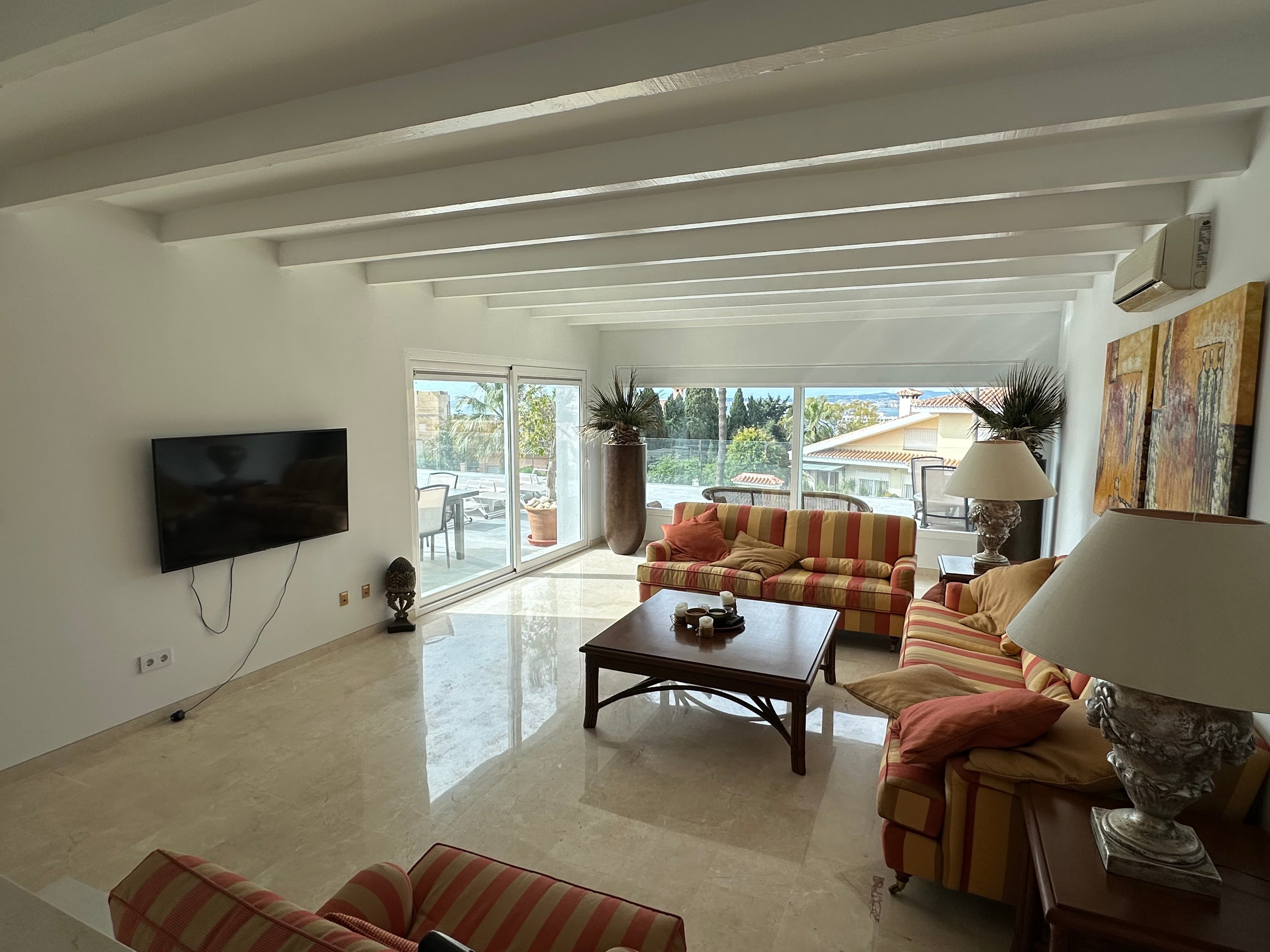 Bright living room with beamed ceiling, striped sofas, and large windows overlooking a sunny patio.