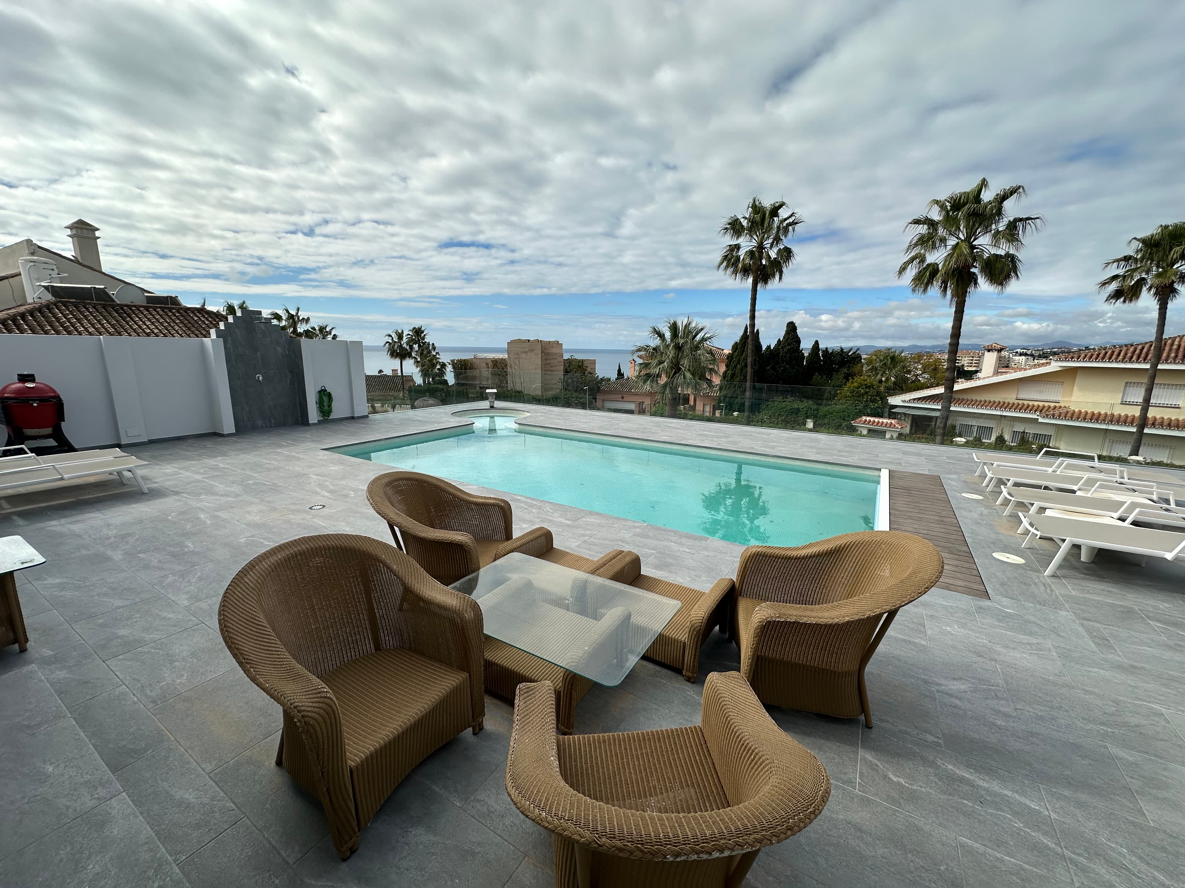 Wicker patio furniture beside a swimming pool with palm trees and distant ocean views.