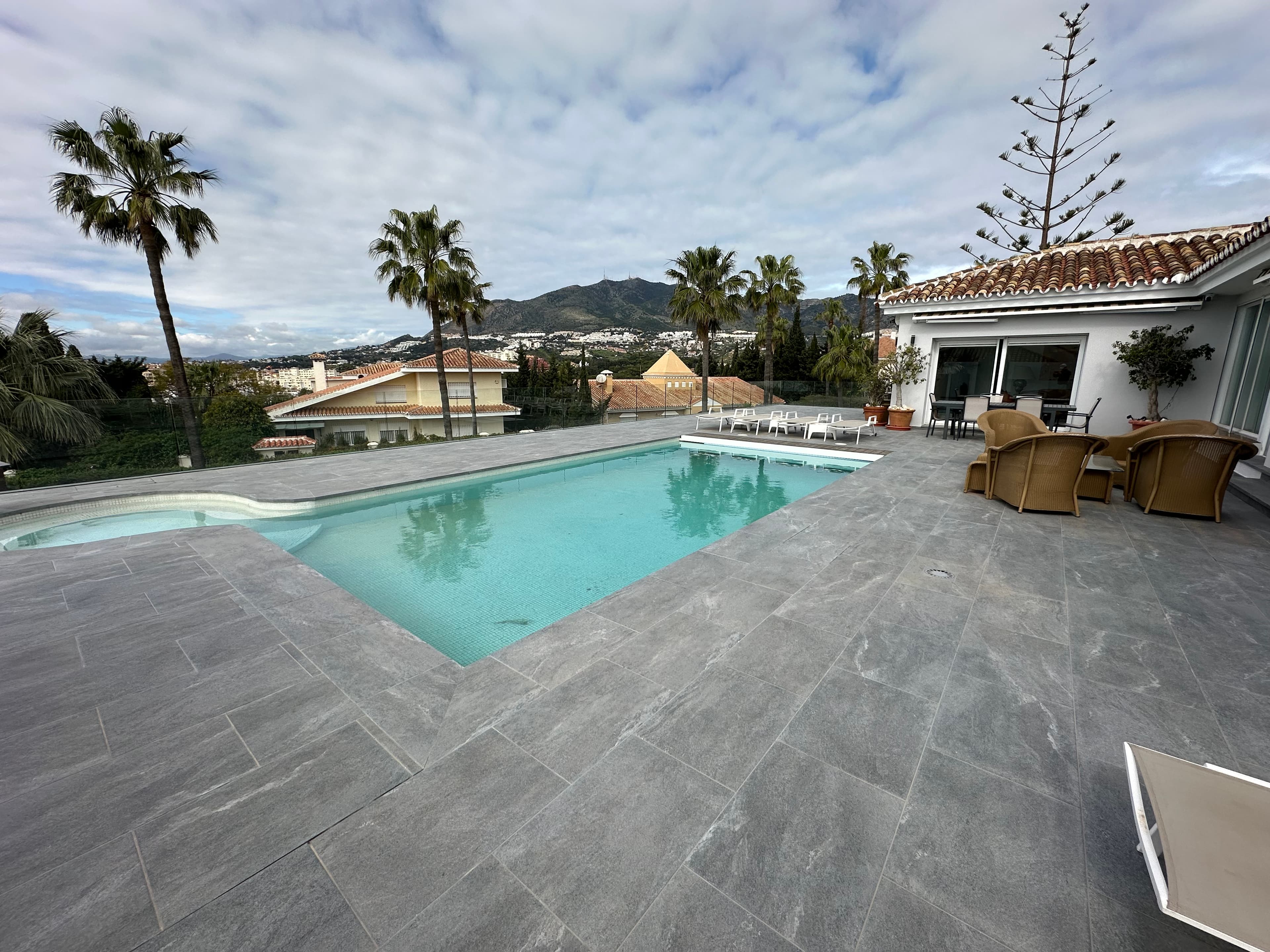 Modern swimming pool on a grey tiled patio overlooking palm trees and distant mountains.