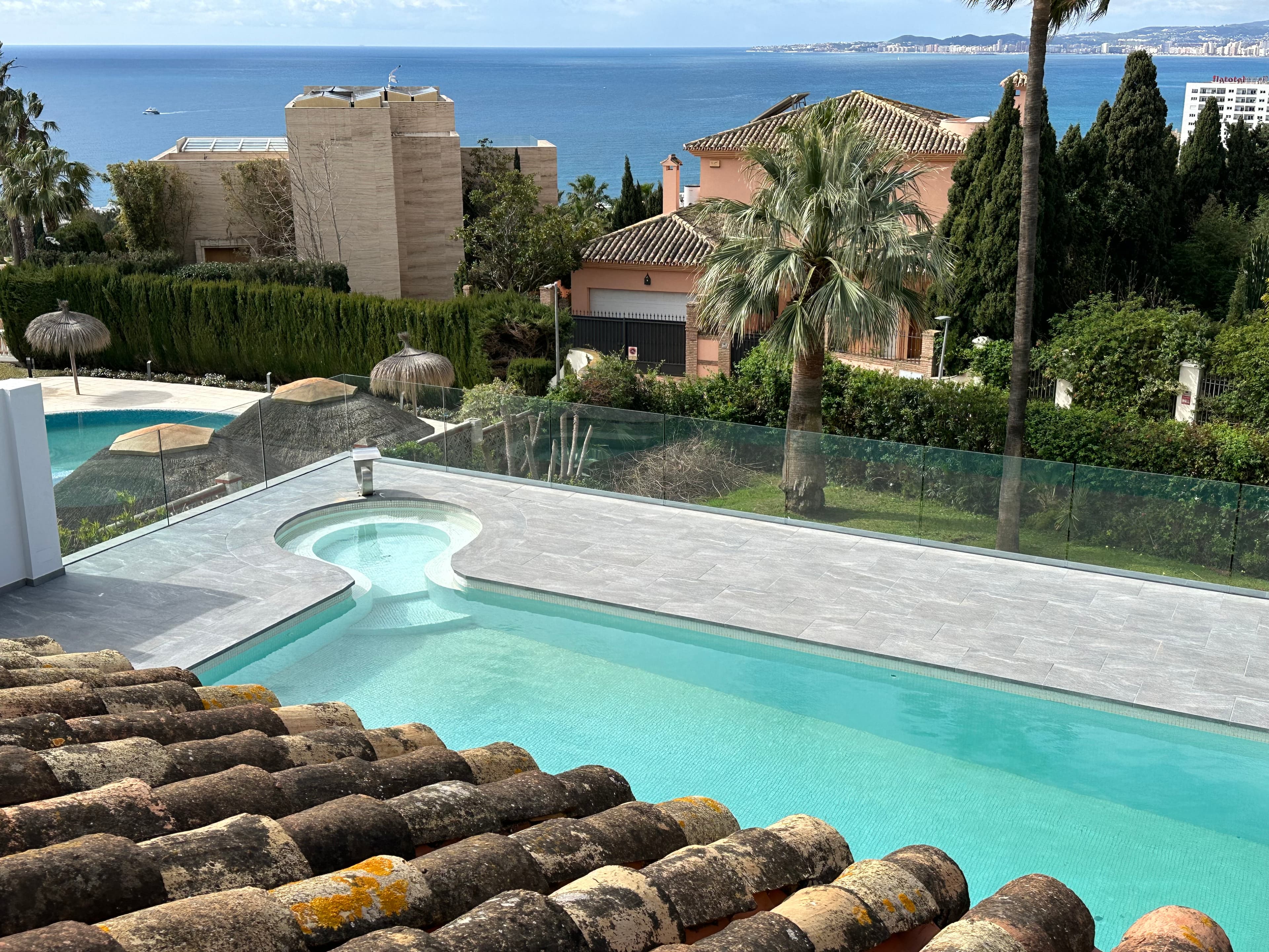 Modern swimming pool on a terrace overlooking Mediterranean houses and the blue sea in the distance.
