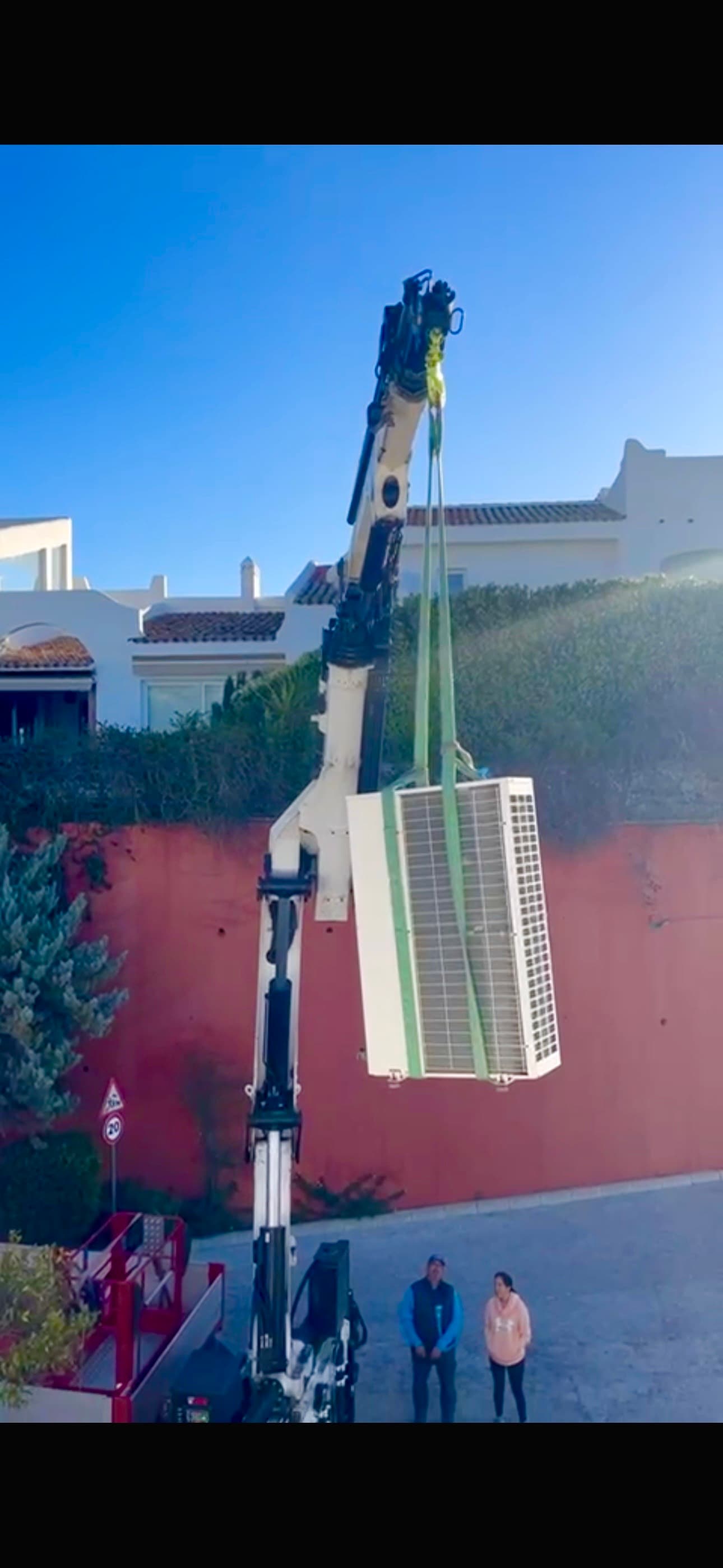 Crane hoisting a large air conditioning unit over a red wall with two people watching.