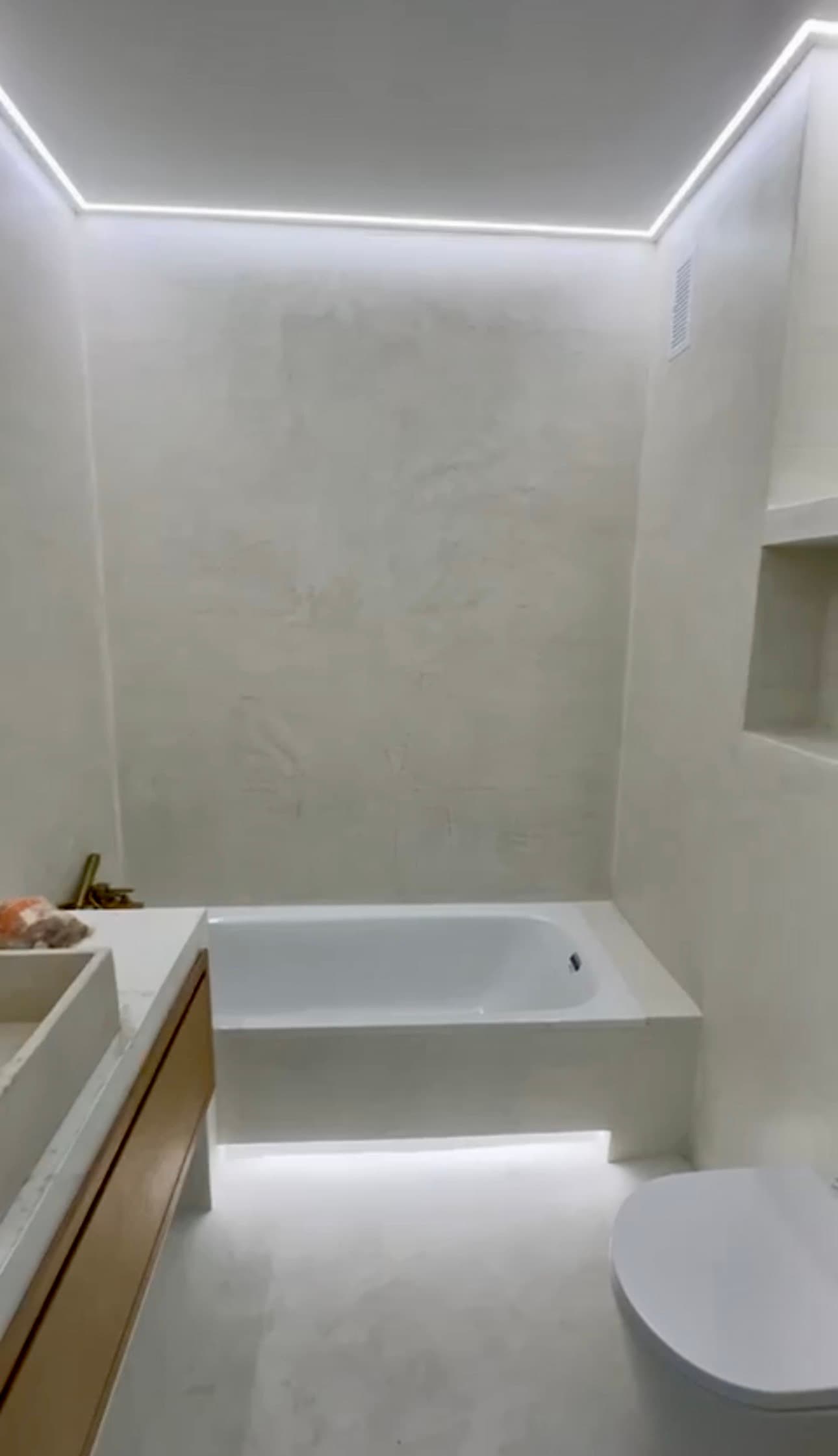 Modern white bathroom with textured walls, a built-in bathtub, and perimeter LED strip lighting.